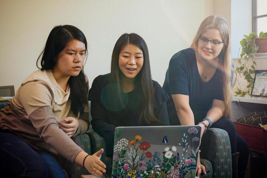 Three students look at a laptop while sitting on a bed.